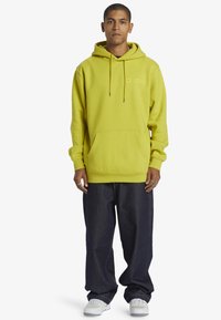 Limegrön bomullshuvtröja med en framficka och snoddar, i kombination med mörk denimjeans och vita sneakers, stående mot en vit bakgrund.