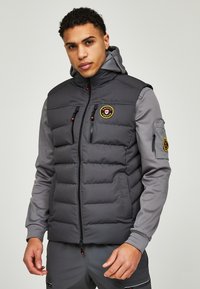 Grijze gewatteerde vest met een ritssluiting aan de voorkant, hoge kraag en kap. Voorzien van een logo patch, ritszakken op de borst en elastische manchetten voor comfort.