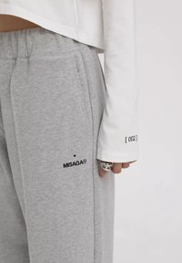 Lichtgrijze sweatpants met een relaxte pasvorm, voorzien van een zwart geborduurd logo "MISAGA®" en een label met de markering "[ 002 ]" aan de zijkant.
