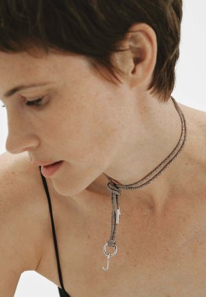 Primer plano de una persona con cabello corto que lleva un top de tirantes finos y un collar de cuerda metálica de doble lazo con un anillo y un pequeño colgante.