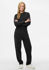 En blonda kvinna står med armarna i kors, klädd i en svart långärmad topp och matchande byxor, tillsammans med vita sneakers mot en vit bakgrund.