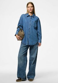 Denim oversized košile s náprsní kapsou, kombinována s širokými džínami. Model drží tašku s tygřím potiskem. Vše v odstínech modré.