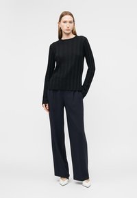 Žena nosiaca čierny rebrovaný dlhý rukáv, námornícke široké nohavice a biele špicaté topánky, stojaca pred jednoduchým pozadím.