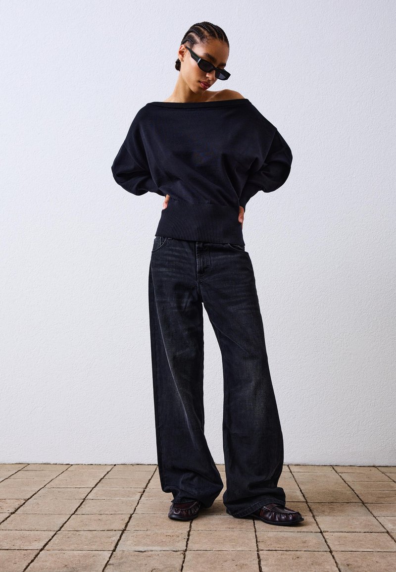 Črna oversized pulover s padlimi rameni in rebrastim robom, v kombinaciji z širokimi temnimi jeans hlačami in črnimi ravnimi čevlji. Teksturni ozadje.