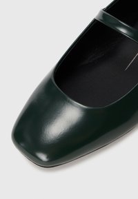 Ballerine in pelle verde con punta quadrata e cinturino alla caviglia. Presentano una finitura lucida e una fodera interna morbida.