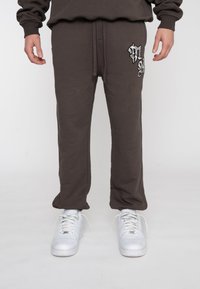 Person iført mørkebrune svedbukser med et sort og hvidt broderet design på overlåret og hvide sneakers.