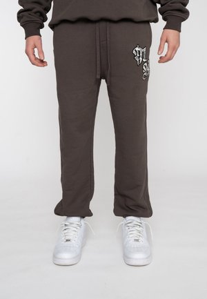 Persona che indossa pantaloni tuta marroni scuri con un design ricamato bianco e nero sulla coscia e sneaker bianche.