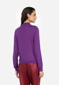 Maglione lunghezza manica viola con collo alto e polsini a costine, abbinato a pantaloni rossi aderenti. Tessuto liscio e design avvolgente.