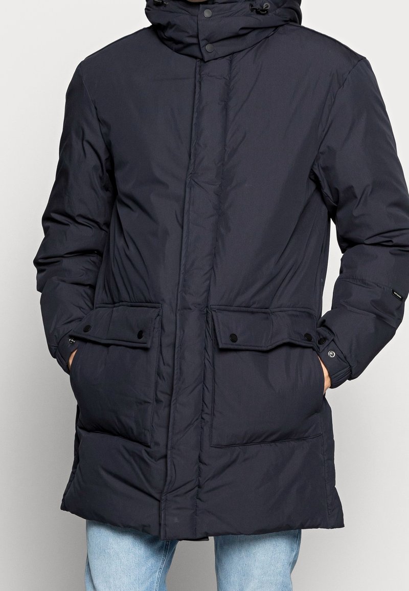 Cappotto imbottito blu marina con cappuccio, ampie tasche con bottoni e chiusura con cerniera. Il materiale è liscio e leggermente imbottito per garantire calore.