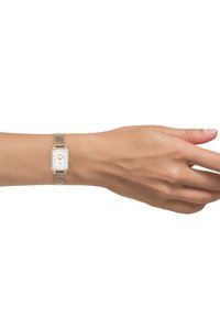 Montre rectangulaire avec un cadran blanc, bracelet en maille dorée et index horaires minimalistes. La montre est portée au poignet d'un teint de peau naturel.