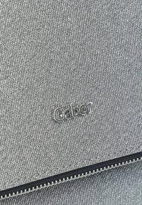 Bolso de tela gris con una superficie texturizada, accesorios plateados y un logo de Gabor. Incluye un detalle de cremallera en la parte inferior.