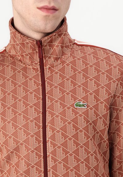 Veste zippée rouge à motifs avec un col haut, présentant un design géométrique et un petit logo de crocodile vert sur la poitrine.