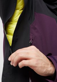 Giacca nera e viola con apertura con zip che rivela un vivace rivestimento giallo. Presenta un design elegante e un tessuto testurizzato. Zip regolabile a mano.