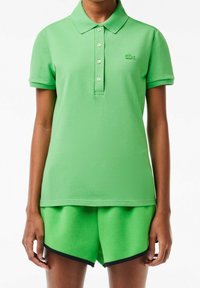 Polo vert avec col et boutons, associé à un short vert assorti avec un tissu texturé et une bordure bleu marine. Logo Lacoste visible.