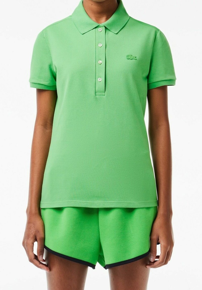 Polo vert avec col et boutons, associé à un short vert assorti avec un tissu texturé et une bordure bleu marine. Logo Lacoste visible.