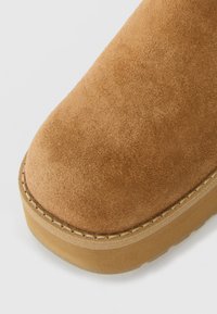 Gros plan sur l'avant d'une botte en daim marron clair avec des coutures visibles et une semelle épaisse en caoutchouc sur un fond clair.