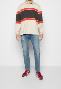 Hombre que lleva una camiseta polo de manga larga a rayas beige, negra y roja, jeans azules claros con desgaste y zapatillas rojas con rayas blancas.
