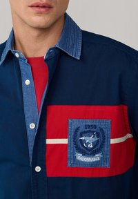 Camicia di cotone con colletto blu, pannello anteriore rosso, logo ricamato e striscia bianca orizzontale. Chiusura frontale con bottoni e design casual.