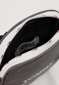 Borsa a tracolla grigia con zip bianca e bordi rosso, bianco e blu, parzialmente aperta che mostra l'interno nero e l'etichetta Tommy Jeans all'interno.