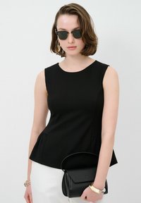 Top nero senza maniche con dettaglio peplum in vita, texture liscia. La modella tiene una piccola borsa nera e indossa occhiali da sole tondi.