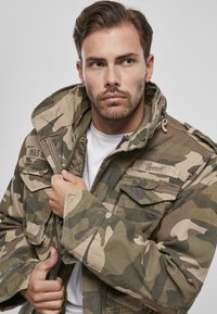 Maskovacia bunda so zipsom vpredu a viacerými vreckami, vyrobená z odolnej látky, v odtieňoch zelenej, hnedej a béžovej.