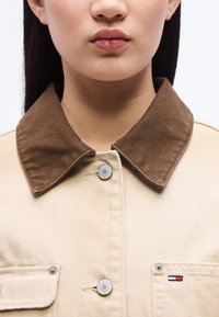 Γυναίκα που φοράει μπεζ μπουφάν με καφέ γιακά, δύο ασημένια κουμπιά και μικρό λογότυπο Tommy Jeans σε σχήμα σημαίας στην τσέπη του στήθους.