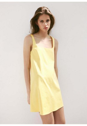 Jeune femme aux longs cheveux bruns portant une robe jaune sans manches, debout devant un fond clair uni, regardant la caméra.