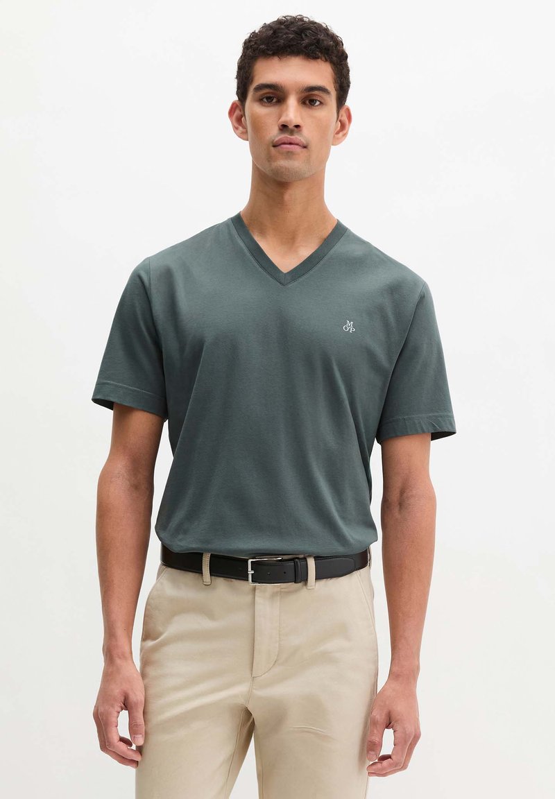 Νέος άνδρας φορώντας σκούρο πράσινο μπλουζάκι V λαιμόκοψη με μικρό λογότυπο, μπεζ παντελόνι και μαύρη ζώνη, στέκεται μπροστά σε απλό λευκό φόντο.