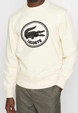 Mann trägt cremefarbenen Langarm-Sweatshirt mit großem schwarzem Lacoste-Krokodil-Logo und Schriftzug, kombiniert mit braunen karierten Hosen.