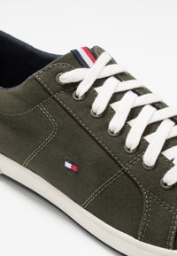 Olivgrön canvas sneakers med vita snörningar, gummisula och en liten broderad logotyp i rött, vitt och blått på sidan.