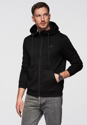 Hombre con sudadera con capucha negra con cremallera y vaqueros grises, mirando hacia un lado con una mano en el bolsillo y reloj en la muñeca.