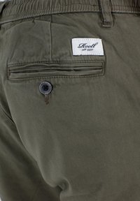 Pantalones de algodón color verde oliva con un bolsillo trasero, con un botón oscuro y una etiqueta de tela blanca que dice "Reell est. 1997". Detalles de costura visibles.