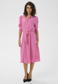 Robe-chemise rose avec manches courtes ballon, boutonnage devant, ceinture à la taille et jupe fluide. Portée avec des chaussures à talons hauts noires. Texture lisse.
