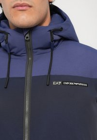 Jūrinės ir mėlynos spalvos kostiumas su pindizuotu gobtuvu, užtrauktuku priekyje, reguliuojamais raišteliais ir EA7 Emporio Armani logotipu ant krūtinės.