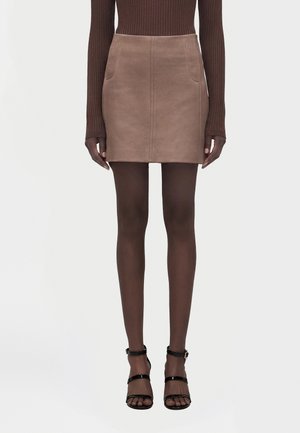 Mannequin à la peau foncée portant un haut marron côtelé à manches longues, une mini-jupe en daim beige, des collants foncés et des talons noirs à brides sur fond blanc.