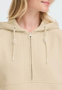 Beige zip-up hoodie featuring a hood, drawstring accents, and a smooth texture. Detailed stitching along the zipper and seams.