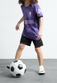 Jersey morado del Liverpool FC con detalles en negro, pantalones cortos negros y zapatos de fútbol color crema. Balón de fútbol con paneles en blanco y negro a los pies.