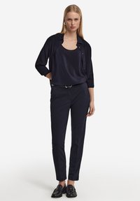 Mörkblå, lätt blus med draperad design, lager under en matchande löst sittande skjorta med knappar. Skräddarsydda mörkblå byxor, slanka svarta loafers.