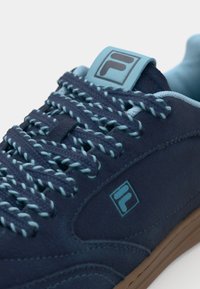 Navy blue sneakers with a textured upper, contrasting light blue laces, branding tag, and brown rubber sole. Features a padded collar.