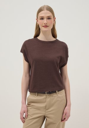 Vrouw met lang blond haar, die gouden oorringen draagt, een los bruin kortarmig topje, beige broek en een zwarte riem, staand tegen een effen achtergrond.