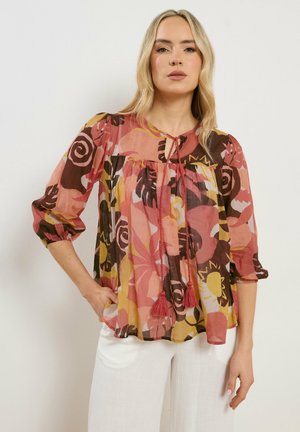 Kobieta w luźnej, kwiecistej bluzce w odcieniach różu, musztardy i brązu, z rękawami do łokcia, zawiązywana frędzlami, w białych spodniach na jednolitym tle.