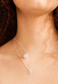 De nek van een vrouw met twee delicate zilveren halskettingen met ronde getextureerde hangers, met krullend haar dat gedeeltelijk zichtbaar is.