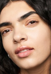 Gros plan sur un visage avec des cheveux foncés et ondulés, une peau douce, des sourcils bien entretenus et une couleur naturelle des lèvres ; met en valeur les traits du visage et la texture.