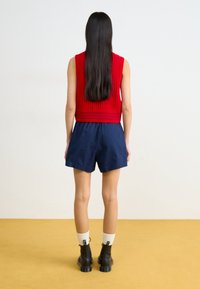 Top sin mangas de punto acanalado rojo con pantalones cortos azul marino, botas negras hasta el tobillo y calcetines blancos, sobre un suelo amarillo y un fondo neutro.