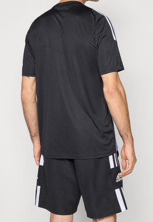 Mężczyzna ubrany w czarną odzież sportową Adidas z białymi paskami na ramionach i szortach, stoi tyłem do kamery.