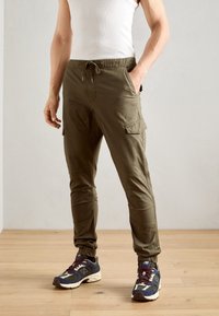 Olijfgroene cargobroek met verstelbare trekkoord tailleband, zijzakken, taps toelopende pijpen en elastische boorden. Combinatie met meerkleurige sneakers.