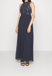 Navy blue maxi dress with a sleeveless design, embellished sheer bodice, and flowing skirt. Worn with black strappy sandals.
