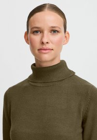 Olivgrön polotröja i mjuk, texturerad tyg med ribbade detaljer vid kragen och axlarna, figurnära silhuett.