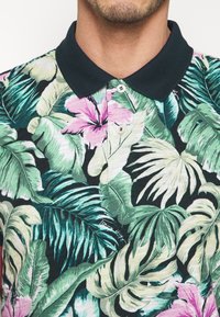 Czarna koszulka polo z tropikalnym zielono-różowym kwiatowym nadrukiem. Posiada czarny kołnierzyk i guziki; materiał jest gładki, bawełniany.