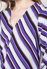 V-neck blouse featuring vertical stripes of purple, blue, brown, and black on a lightweight fabric. Smooth texture with a wrap design.